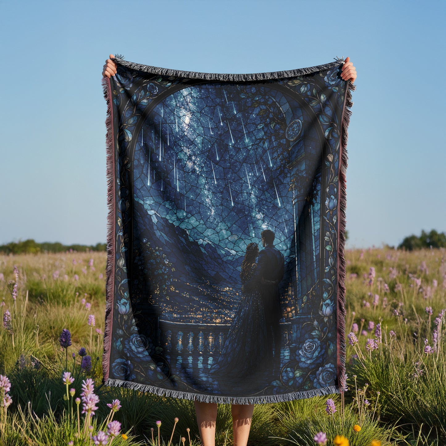 A person is holding up a large, colorful blanket with a picture of a couple standing in front of a scenic landscape, including mountains and a starry sky.
