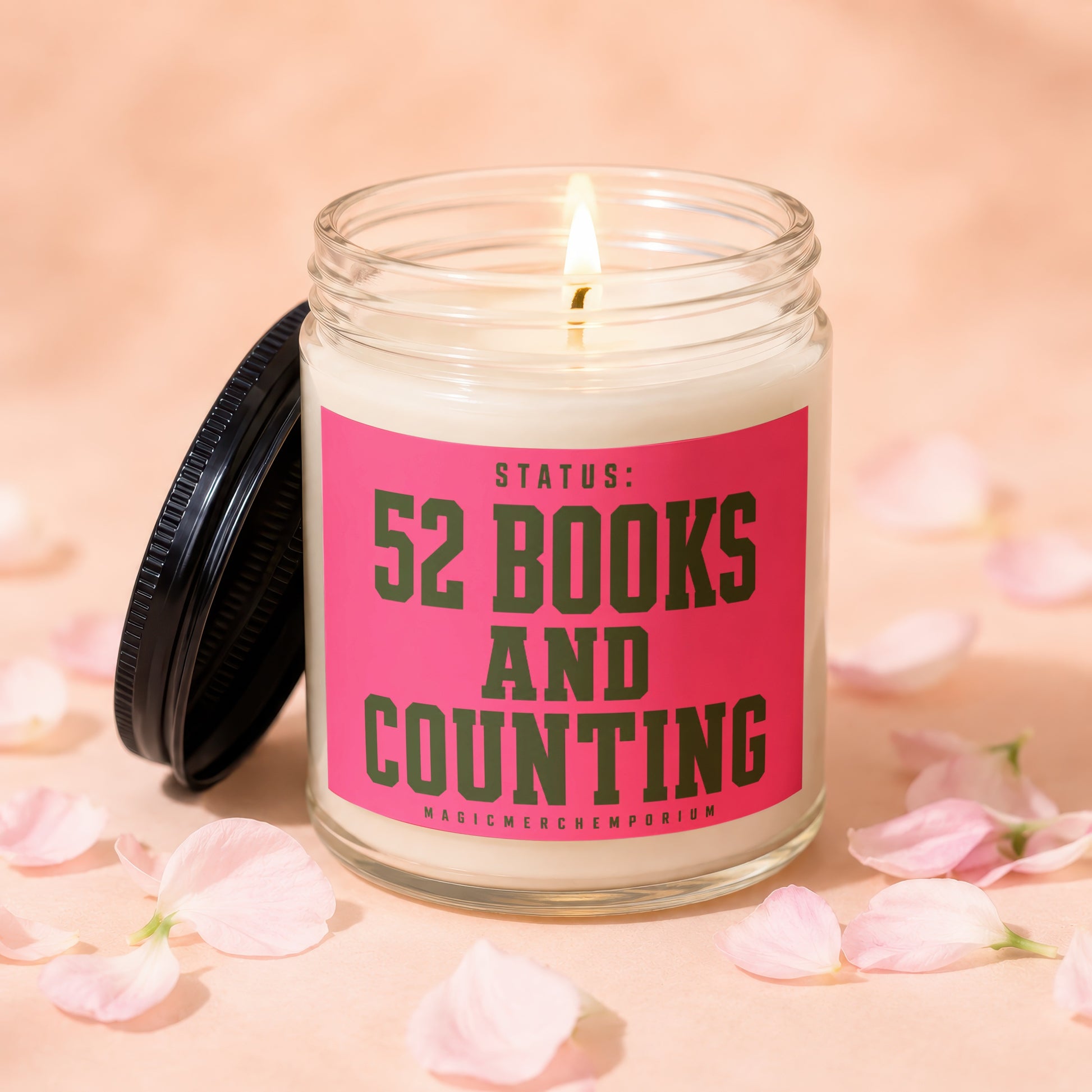 Jar candle with lit wick, featuring pink label with green text "STATUS: 52 BOOKS AND COUNTING".