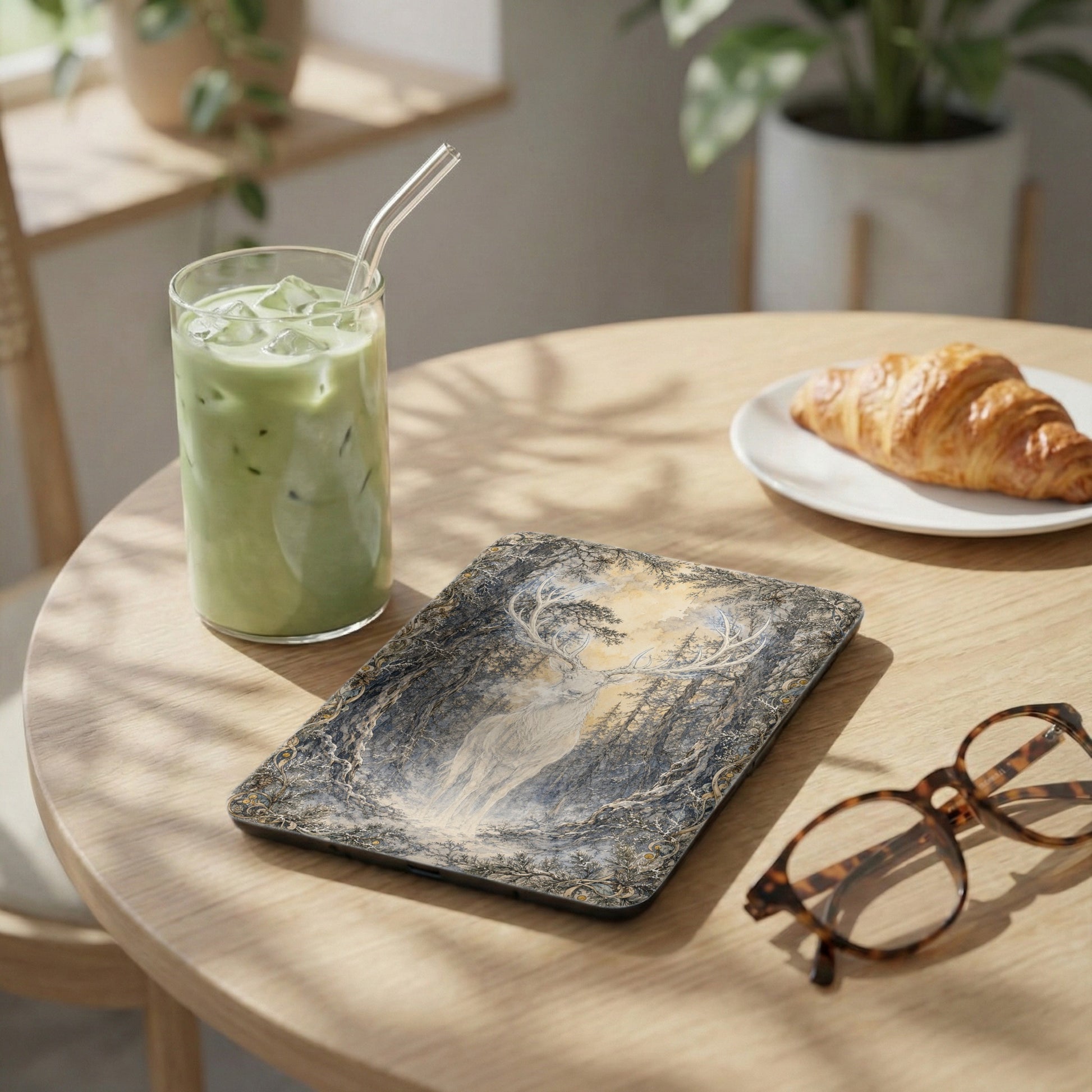 Device with white stag in forest artwork, beside matcha drink and croissant.