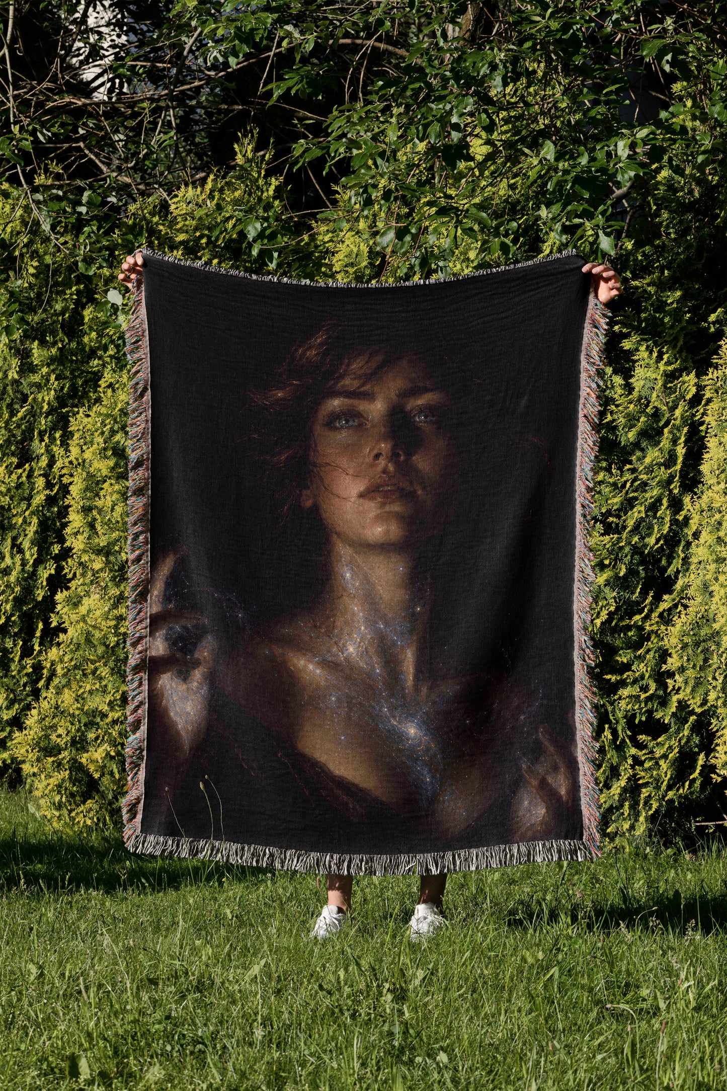 A person is holding a large black blanket with a portrait of a woman on it, standing in a grassy area with trees in the background.