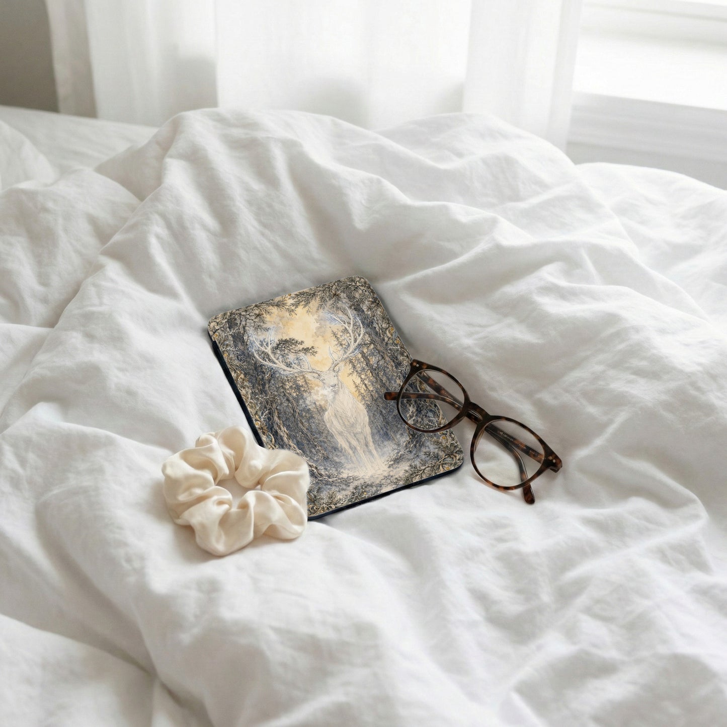 Book with a majestic stag illustration, glasses, and scrunchie on rumpled white bedding.