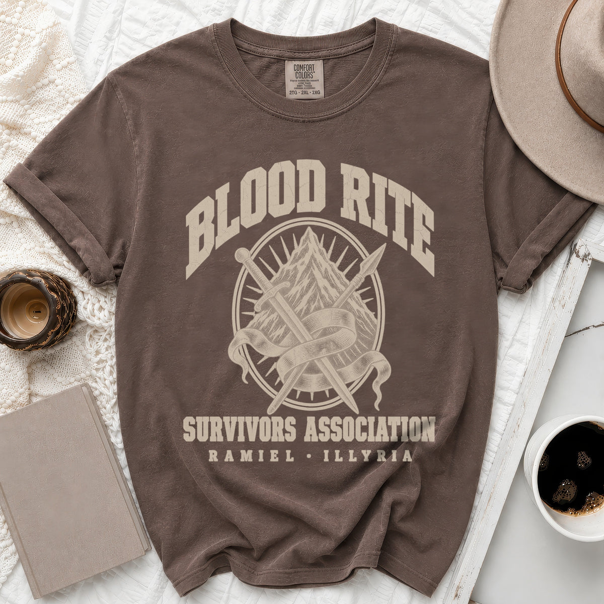A brown crewneck t-shirt featuring the text "Blood Rite Survivors Association Ramiel Illyria" and a mountain graphic with crossed swords.