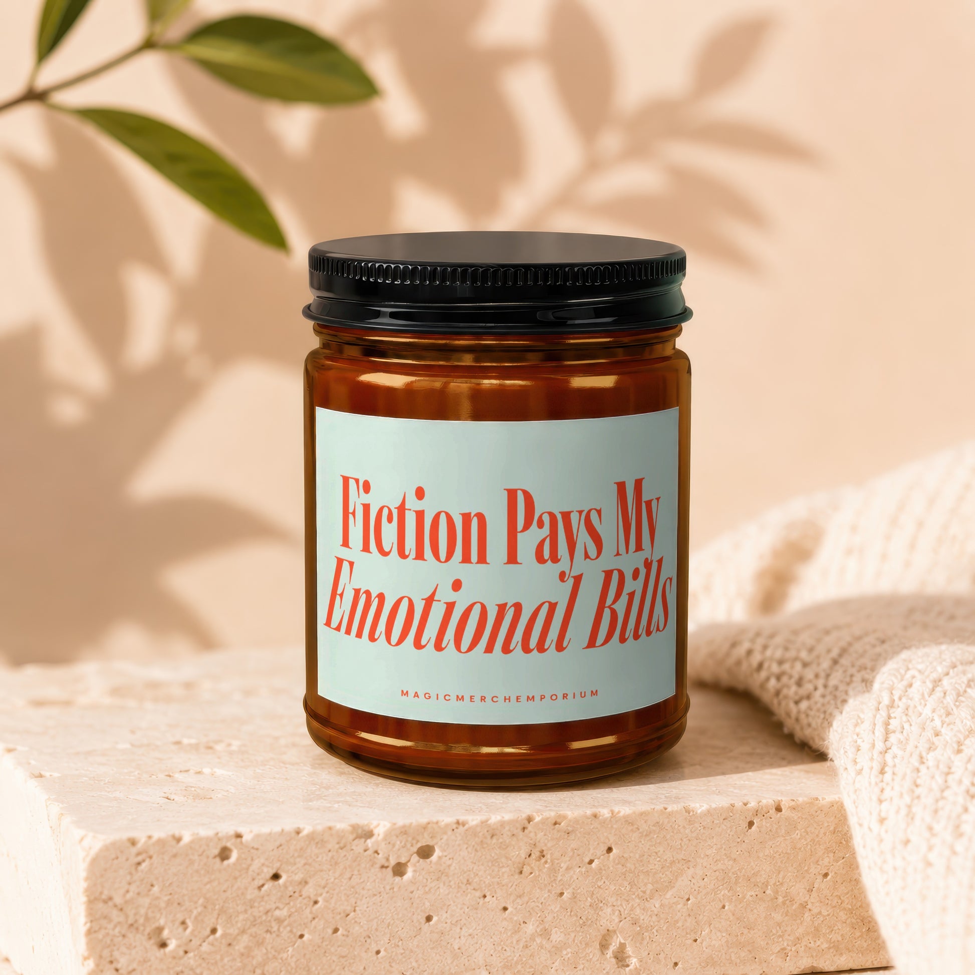 Amber jar candle with text "Fiction Pays My Emotional Bills" on a textured block.