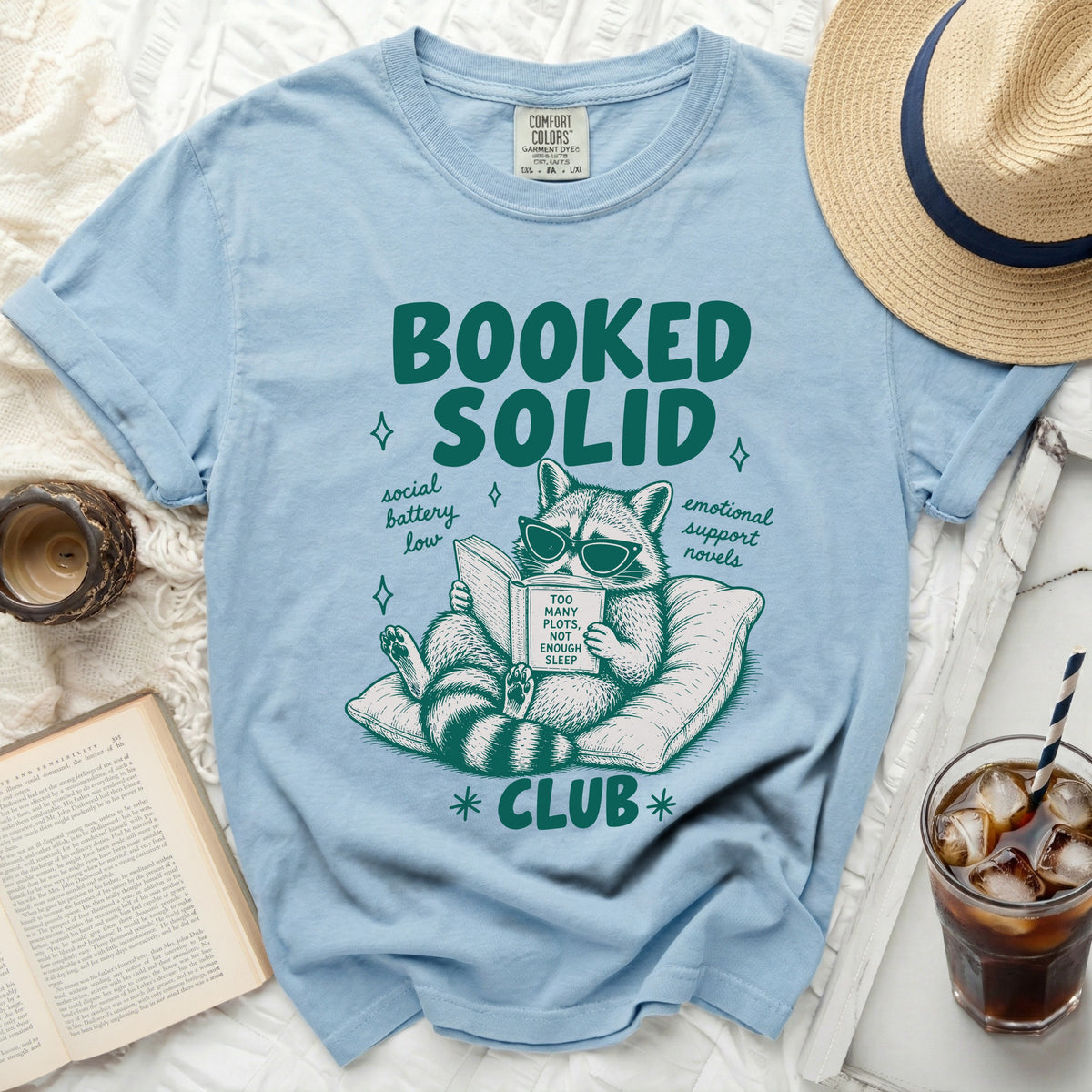 Light blue t-shirt with graphic of raccoon reading book titled "Too Many Plots, Not Enough Sleep".