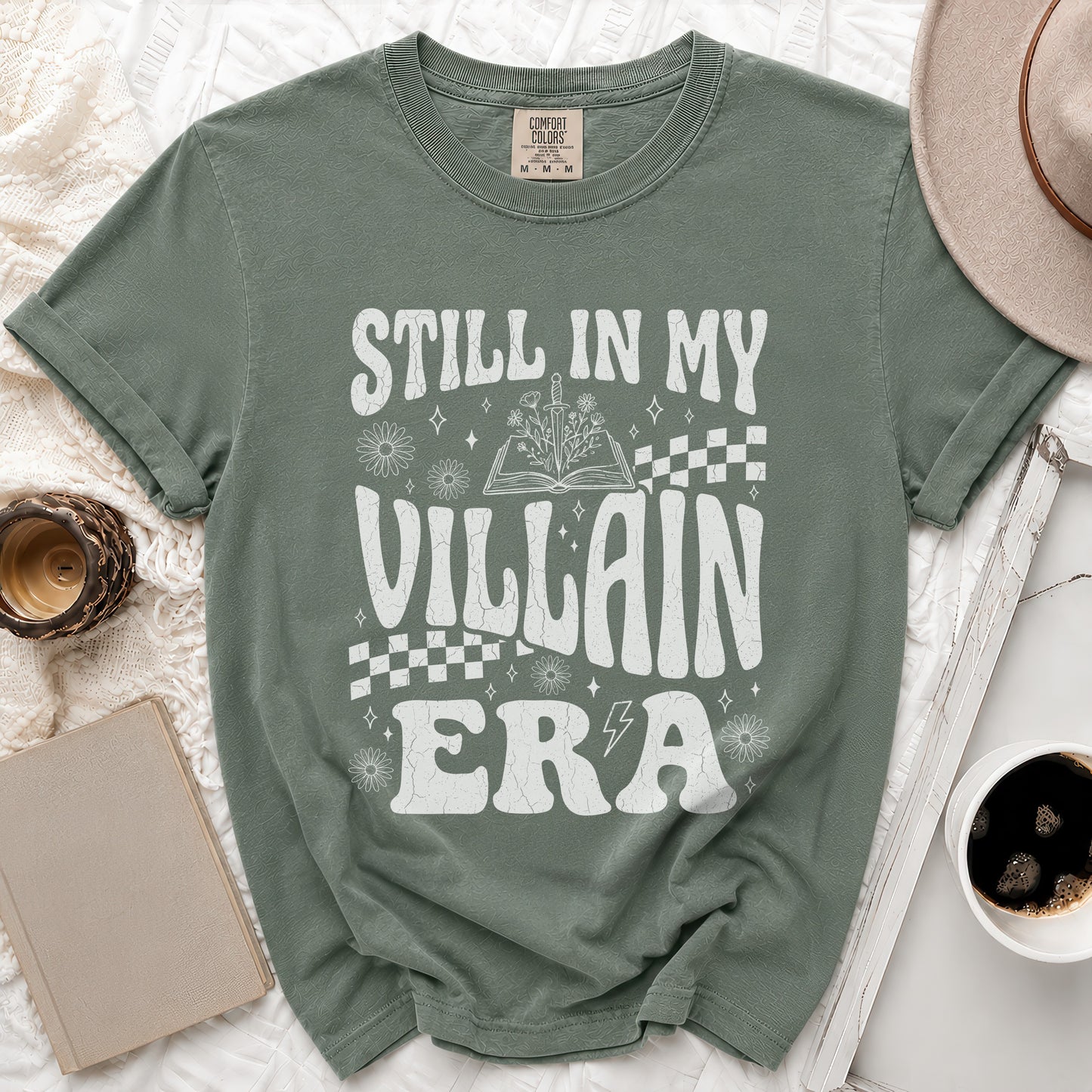 Green t-shirt with white text "Still in My Villain Era" and floral illustrations.