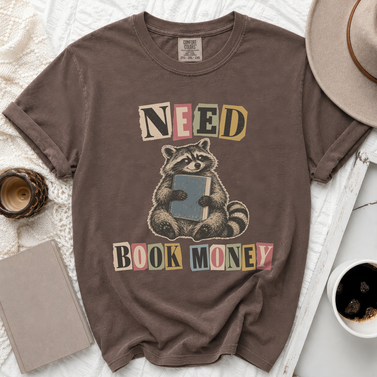 Brown t-shirt with a raccoon holding a book, text reads "NEED BOOK MONEY".