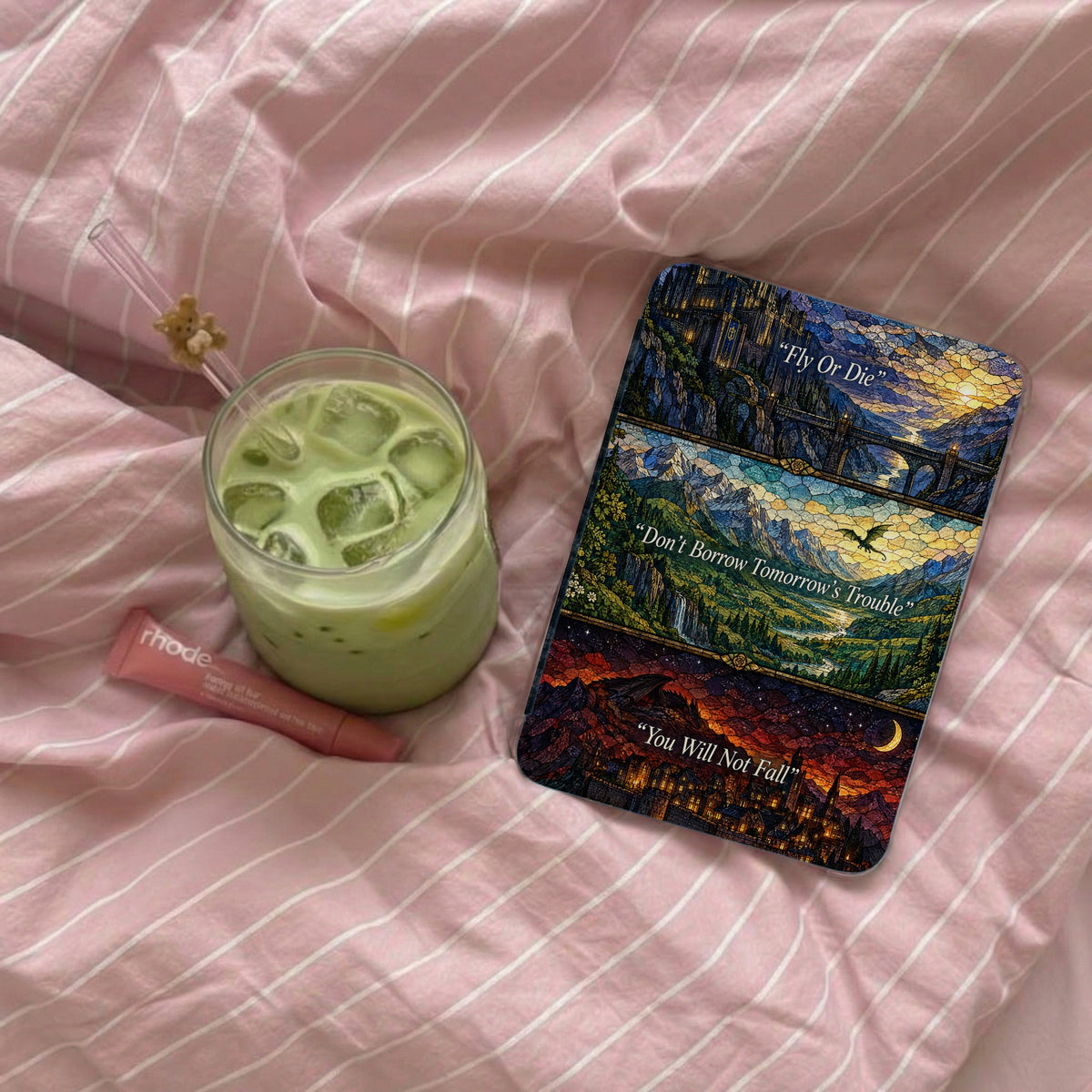An e-reader with a stained glass art skin rests on pink striped bedding next to an iced matcha latte and lip balm.