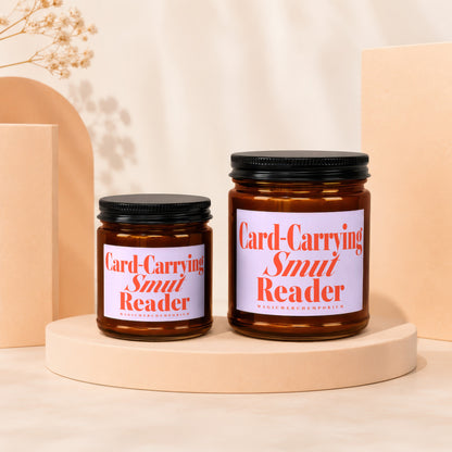 Two glass candles with black lids displaying "Card-Carrying Smut Reader" in orange text.