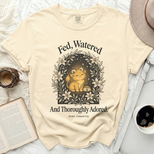 Cream t-shirt with a graphic of a cat surrounded by flowers and text: "Fed, Watered And Thoroughly Adored."