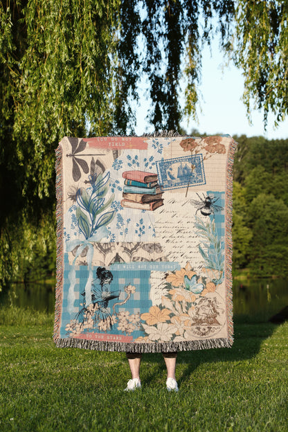 A person is standing in a grassy area, holding a colorful blanket with various illustrations and designs, including a bicycle, flowers, and a bird.