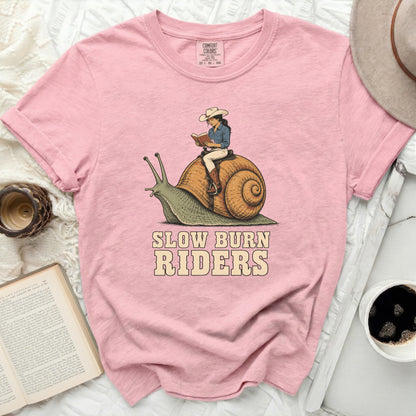 Pink t-shirt with a graphic of a woman riding a snail and text "SLOW BURN RIDERS".