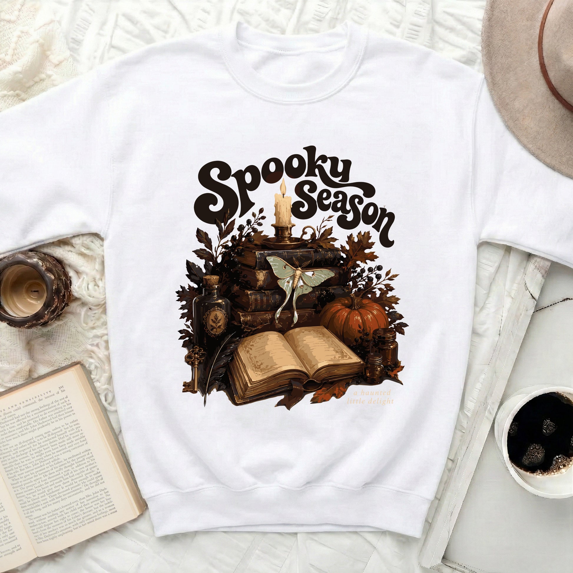 White sweatshirt with "Spooky Season" design featuring books, moth, candle, and pumpkin.