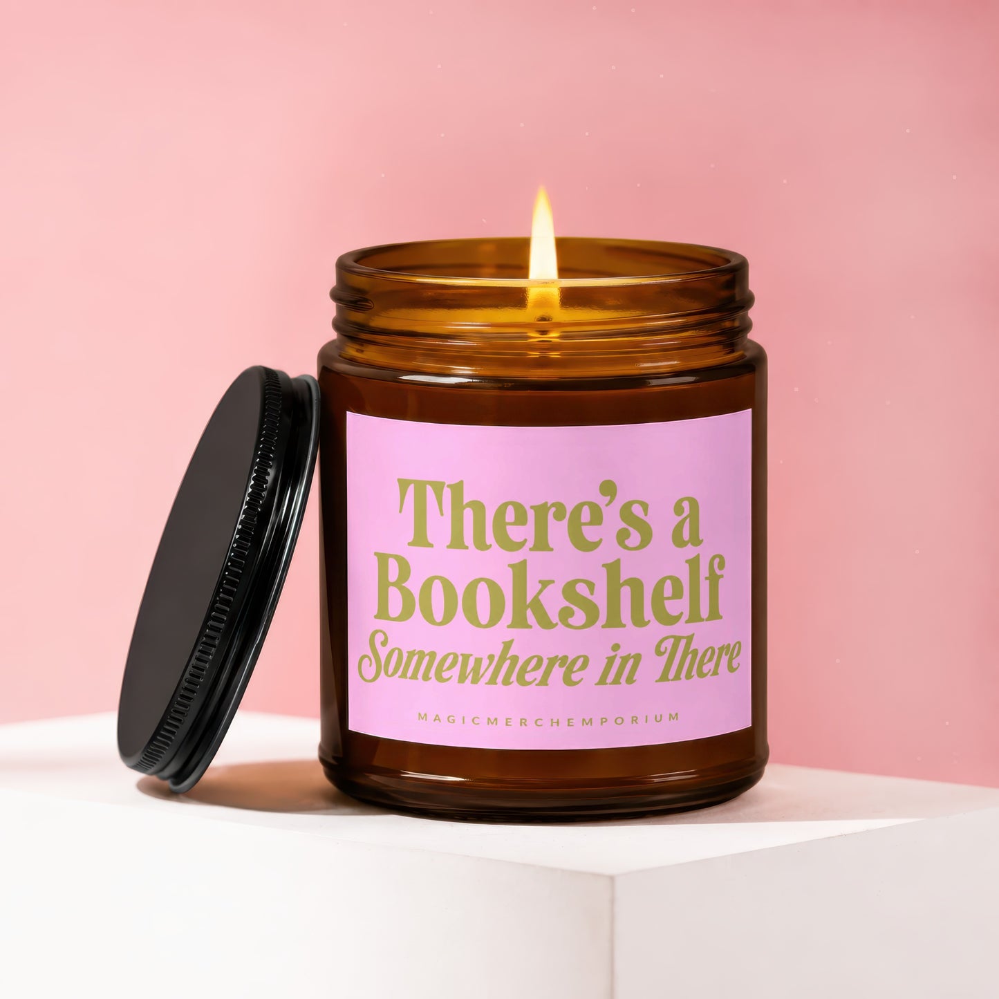 Amber candle with lit flame and pink label reading "There's a Bookshelf Somewhere in There".