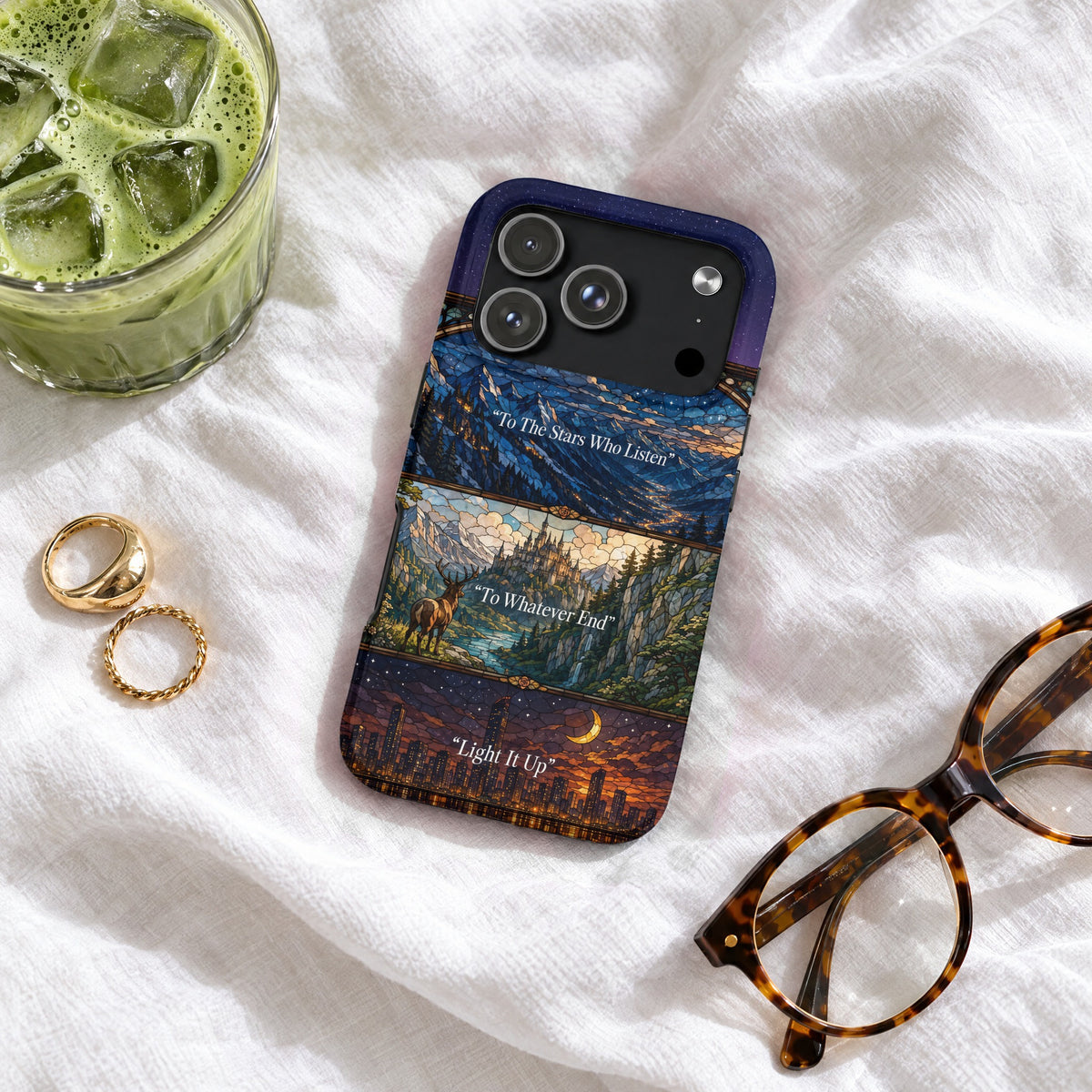 A dark fantasy stained glass phone case displaying quotes sits on a white fabric surface with glasses and iced matcha.