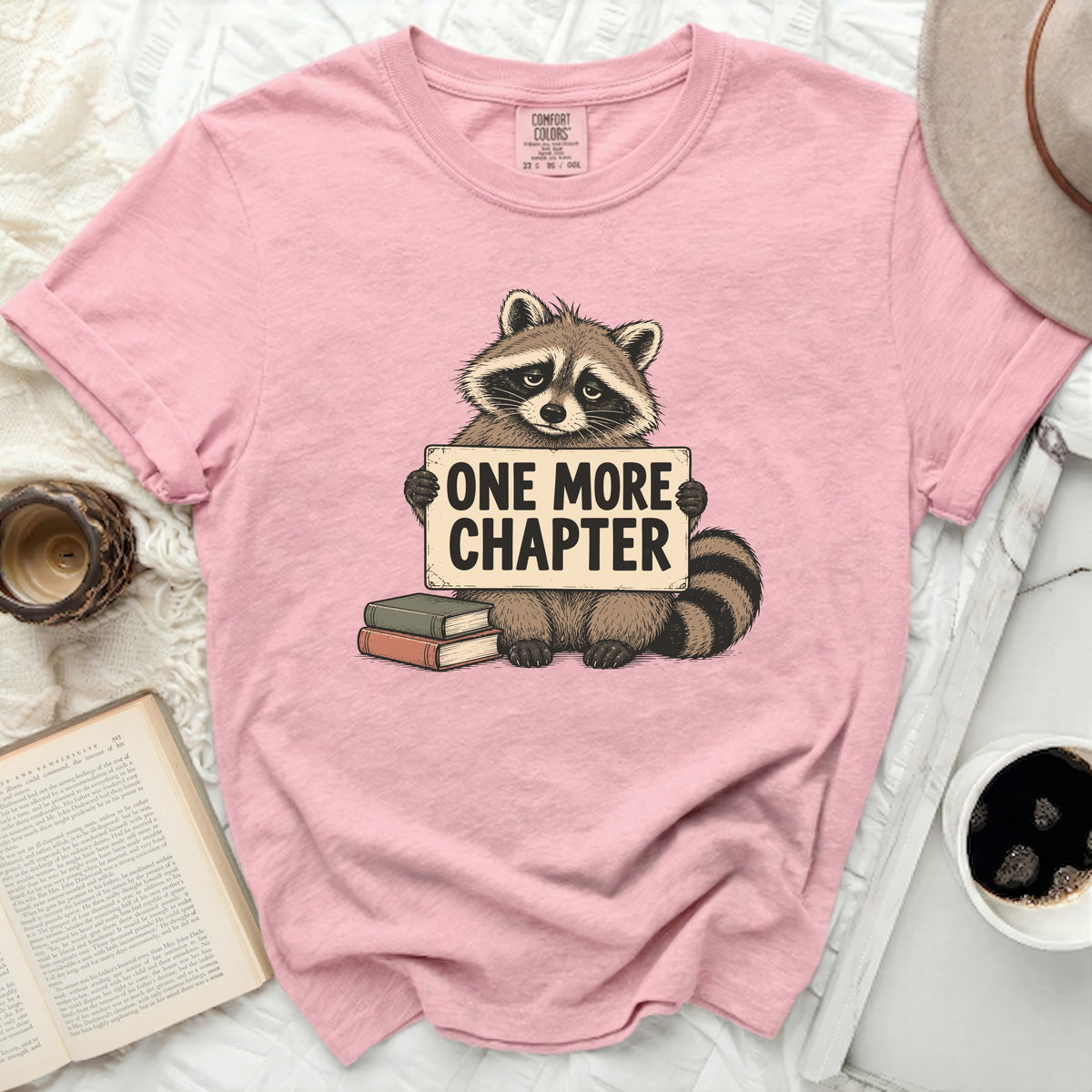 Pink t-shirt with a raccoon holding a sign that says "ONE MORE CHAPTER".