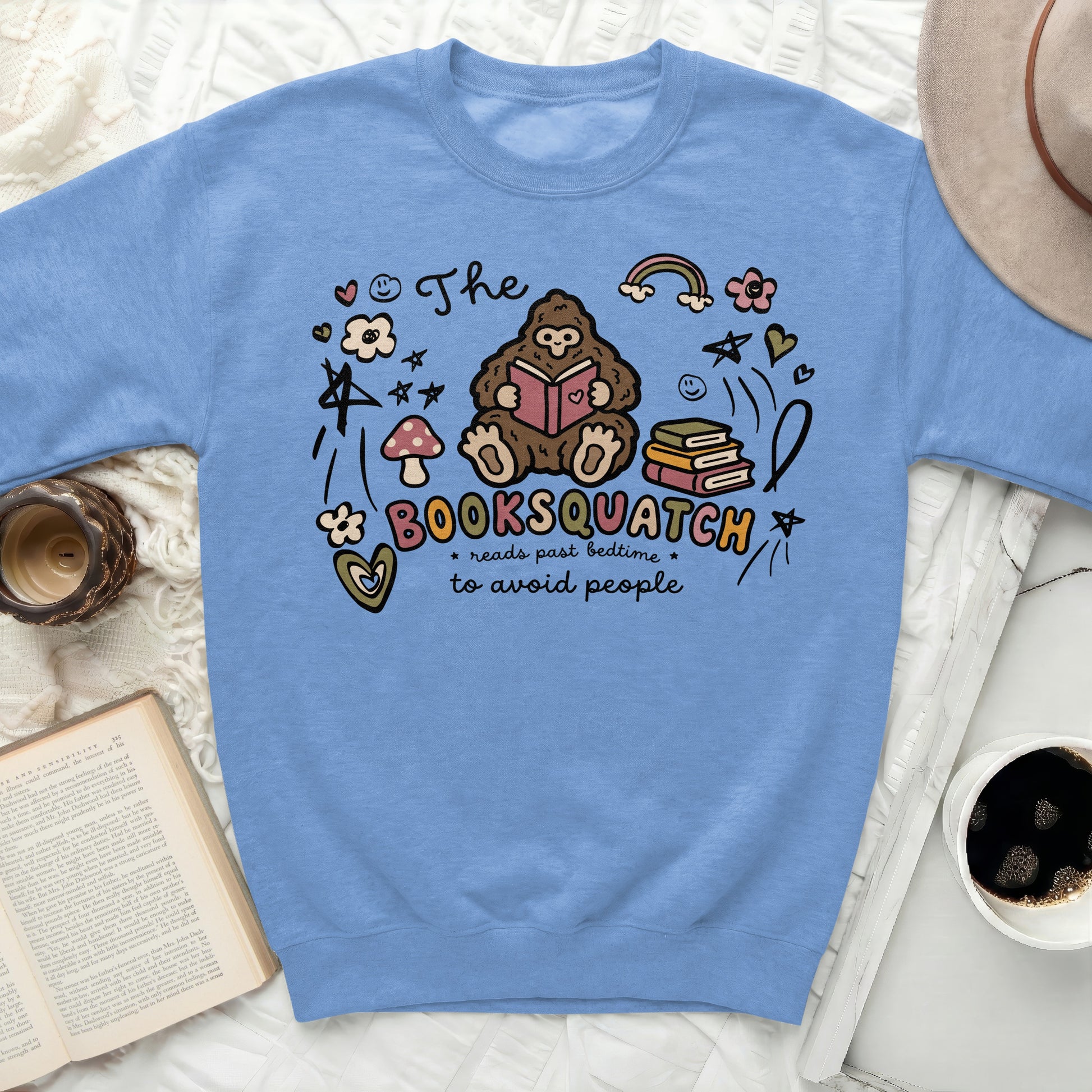 Blue sweatshirt with graphic of Booksquatch reading, books, mushroom, rainbow, and stars.