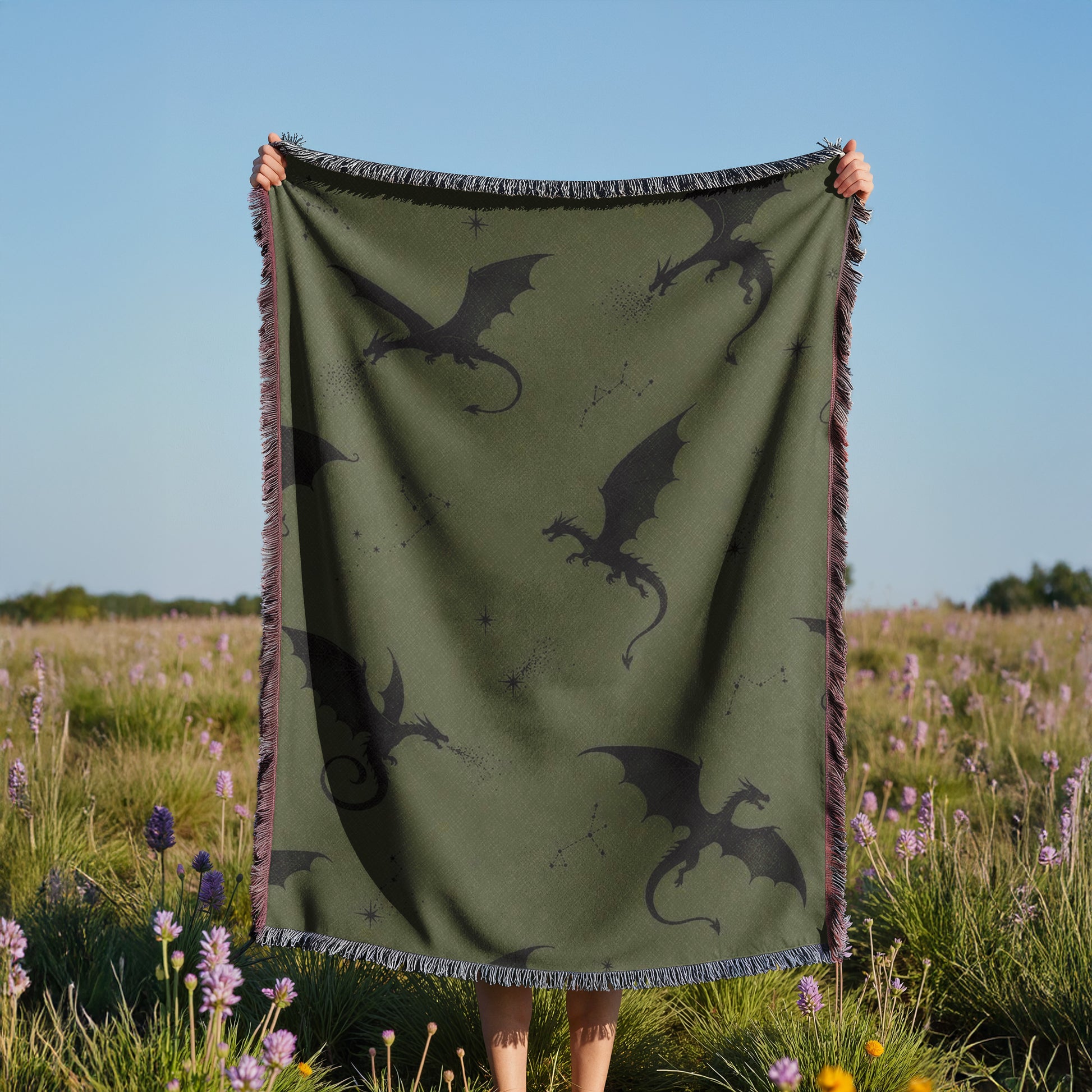 A person is holding up a green blanket with black dragon silhouettes on it, standing in a field with purple flowers.