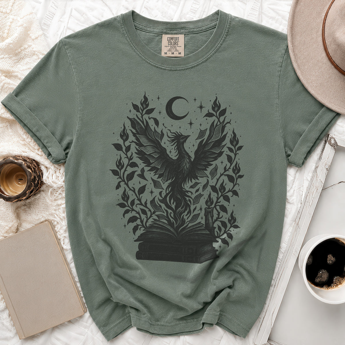 Muted green t-shirt featuring a black graphic of a rising phoenix, stacked books, moon, and foliage.