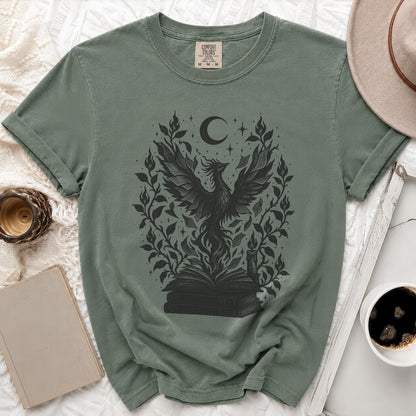 Muted green t-shirt featuring a black graphic of a rising phoenix, stacked books, moon, and foliage.