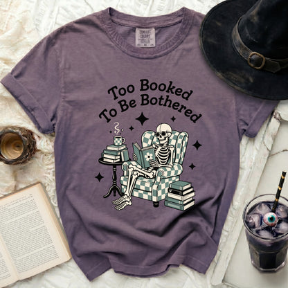 Purple graphic tee with skeleton reading a book in a checkered armchair.