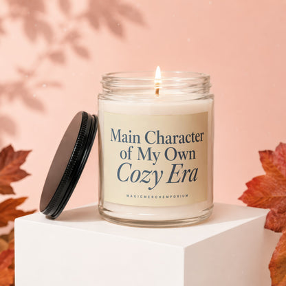 Lit candle with label "Main Character of My Own Cozy Era" on a white cube.