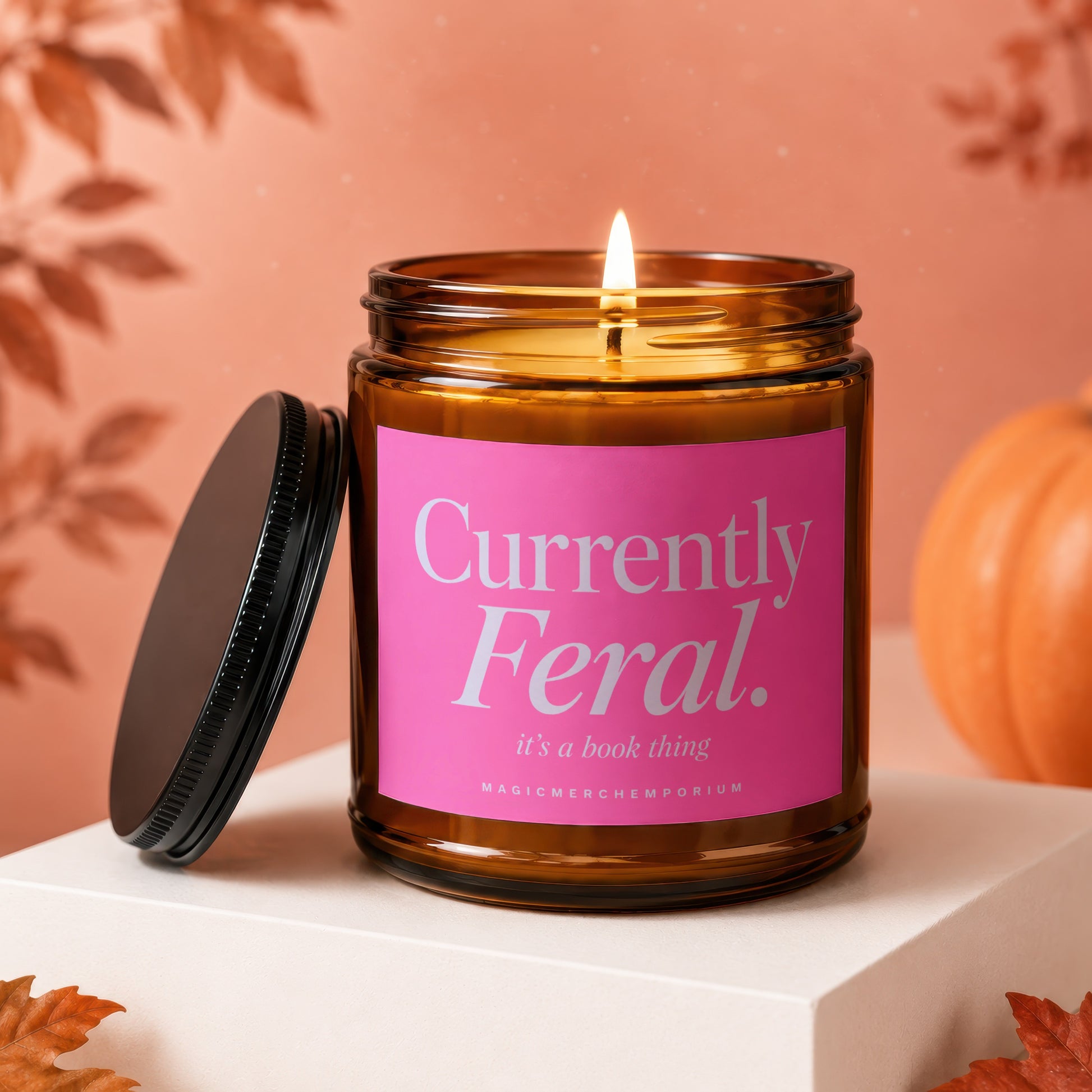 Amber jar candle with pink label reading "Currently Feral. it's a book thing.", lit wick.