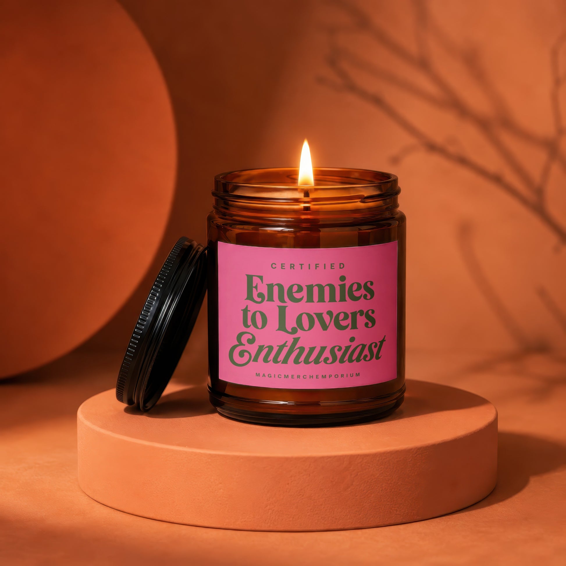 Amber jar candle with lit wick and pink label reading "Certified Enemies to Lovers Enthusiast".