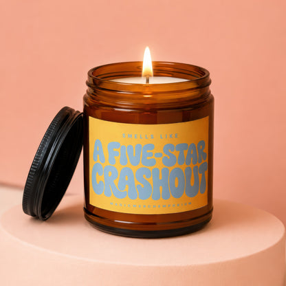 Amber glass candle with lit wick and black lid, label reads "Smells Like A Five-Star Crashout".