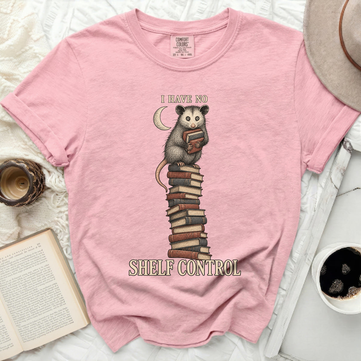 Pink t-shirt with graphic of an opossum holding books on a stack, text "I HAVE NO SHELF CONTROL".