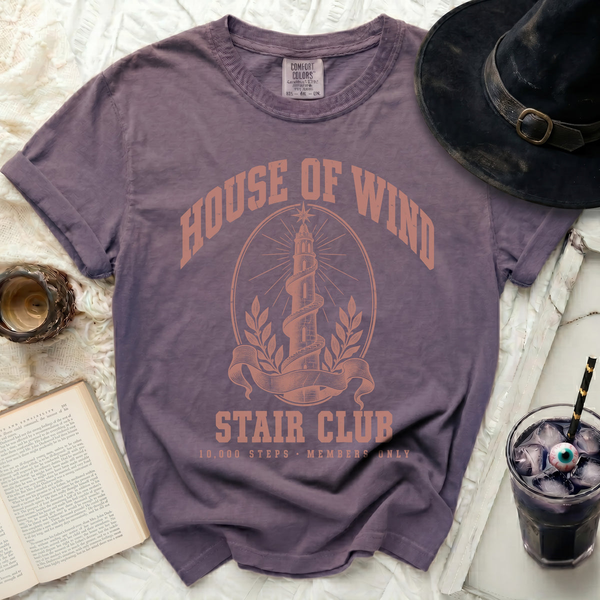 A purple crewneck t-shirt features a peach-colored graphic reading "House of Wind Stair Club" with a spiraled tower illustration.