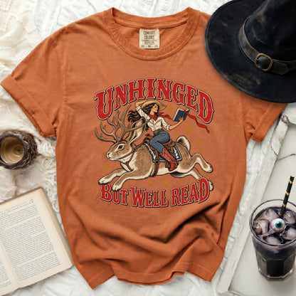 Orange t-shirt with "Unhinged But Well Read" graphic of cowgirl riding jackalope.