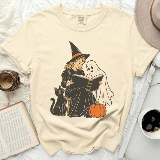 Witch, ghost, cat, and pumpkin reading a book on a cream t-shirt.