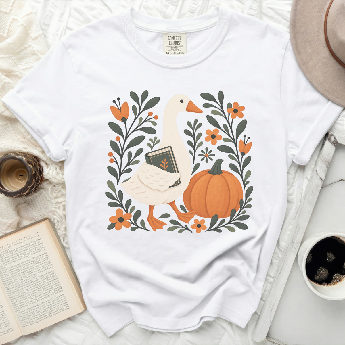 White t-shirt with a goose holding a book surrounded by pumpkins and flowers.