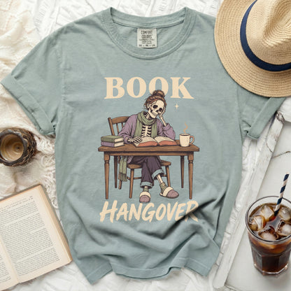 Skeleton reading at a desk with text "BOOK HANGOVER" on a t-shirt.