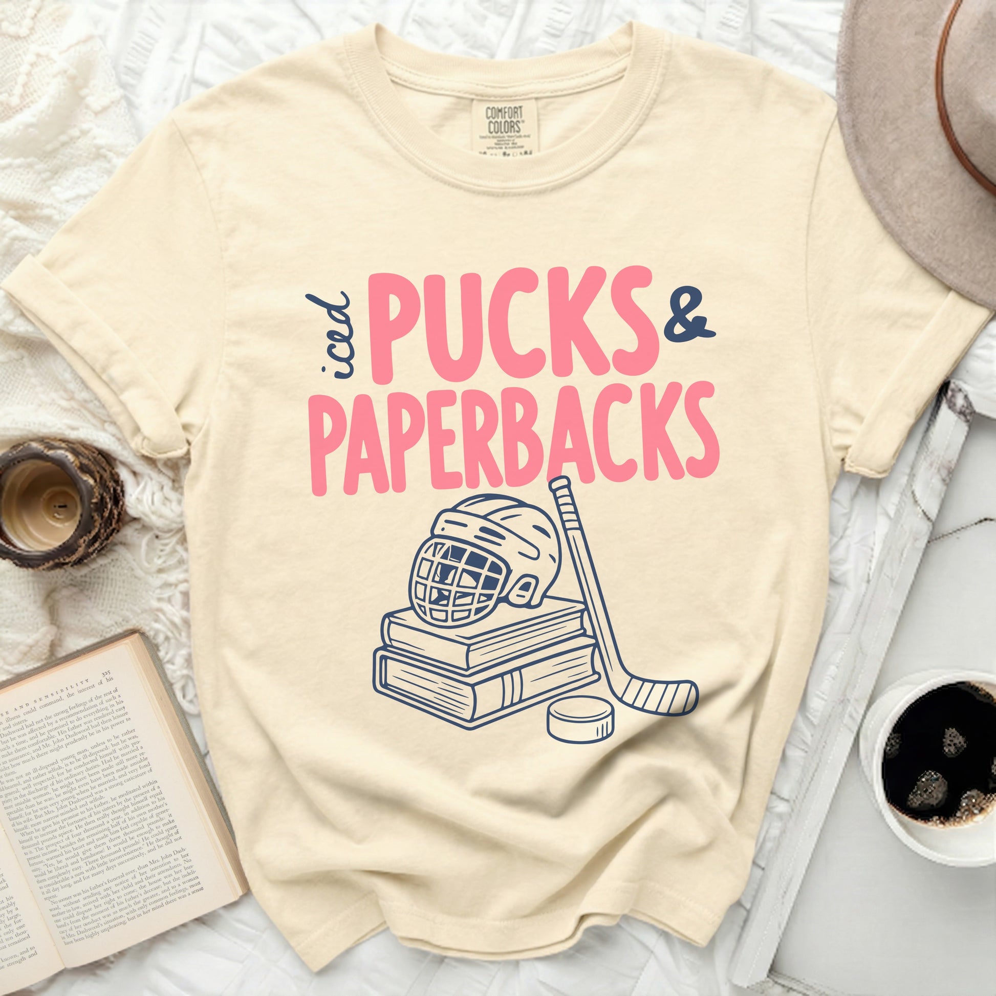 Cream t-shirt with "Pucks & Paperbacks" text, hockey puck, stick, and helmet.