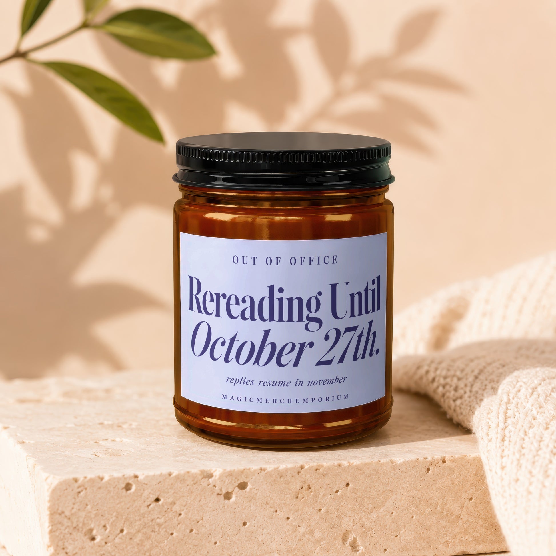 An amber glass candle jar with a black lid sits on a travertine block, featuring a label that reads "Rereading Until October 27th."