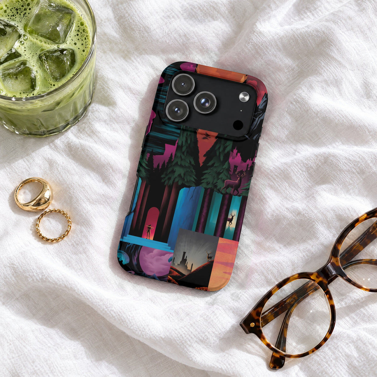 An artistic phone case with a magical forest illustration lies on white fabric near iced matcha, rings, and glasses.