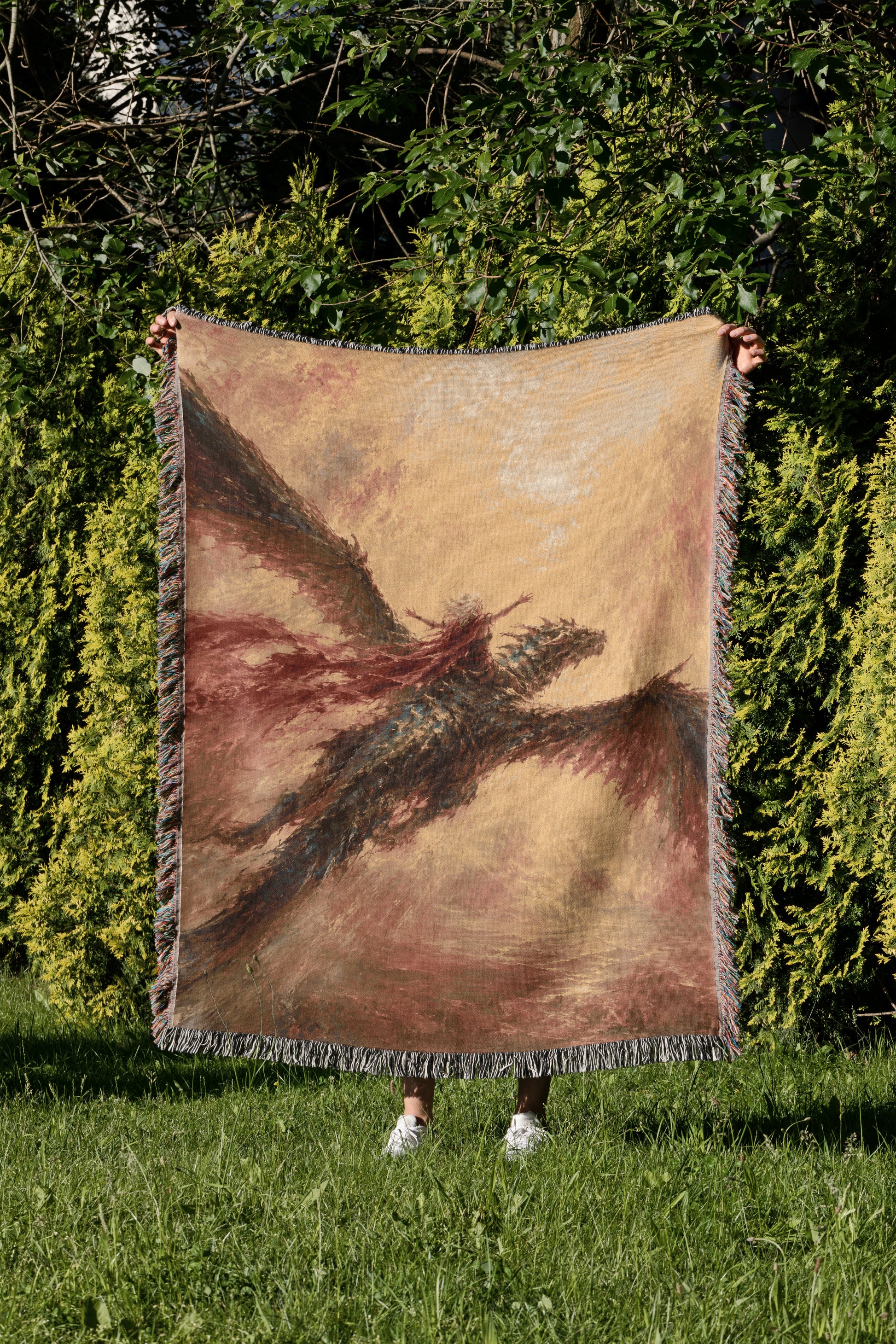A person is holding up a large woven blanket with a dragon design on it, standing in a grassy area with trees in the background.