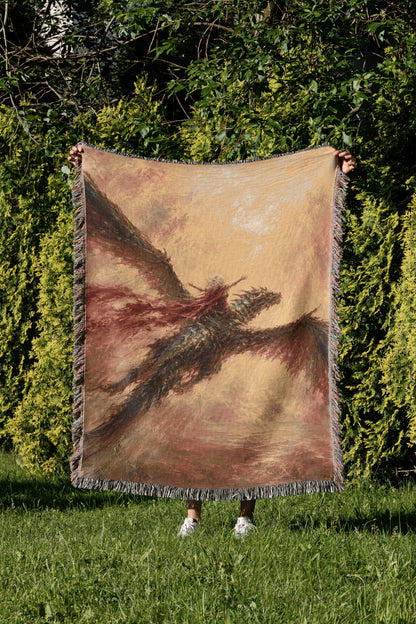 A person is holding up a large woven blanket with a dragon design on it, standing in a grassy area with trees in the background.