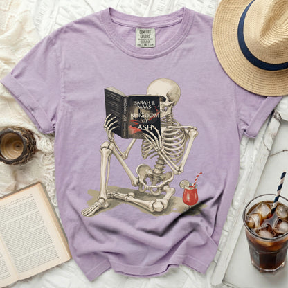 Skeleton reading "Kingdom of Ash" by Sarah J. Maas book with a drink.