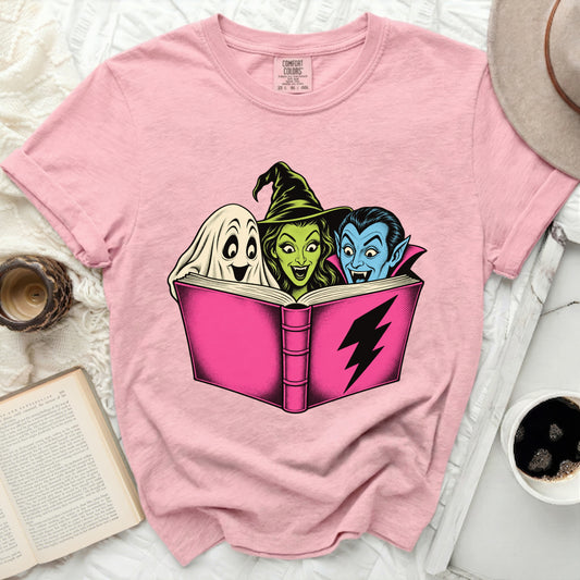 Pink t-shirt with a graphic of a ghost, witch, and vampire reading a book.
