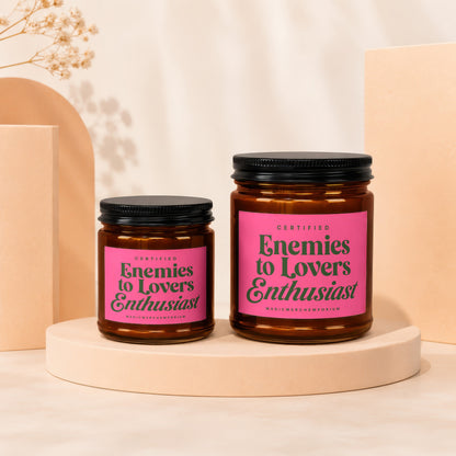 Two amber glass candles with pink labels reading "Certified Enemies to Lovers Enthusiast".