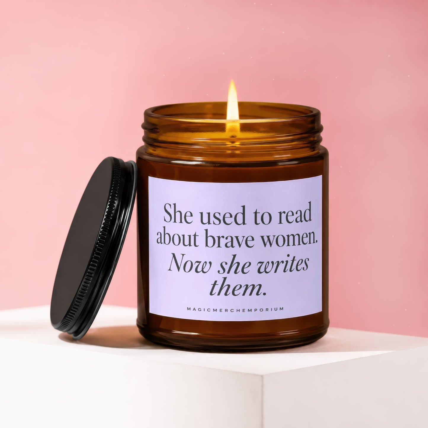 Amber glass candle with lit flame and text "She used to read about brave women. Now she writes them."