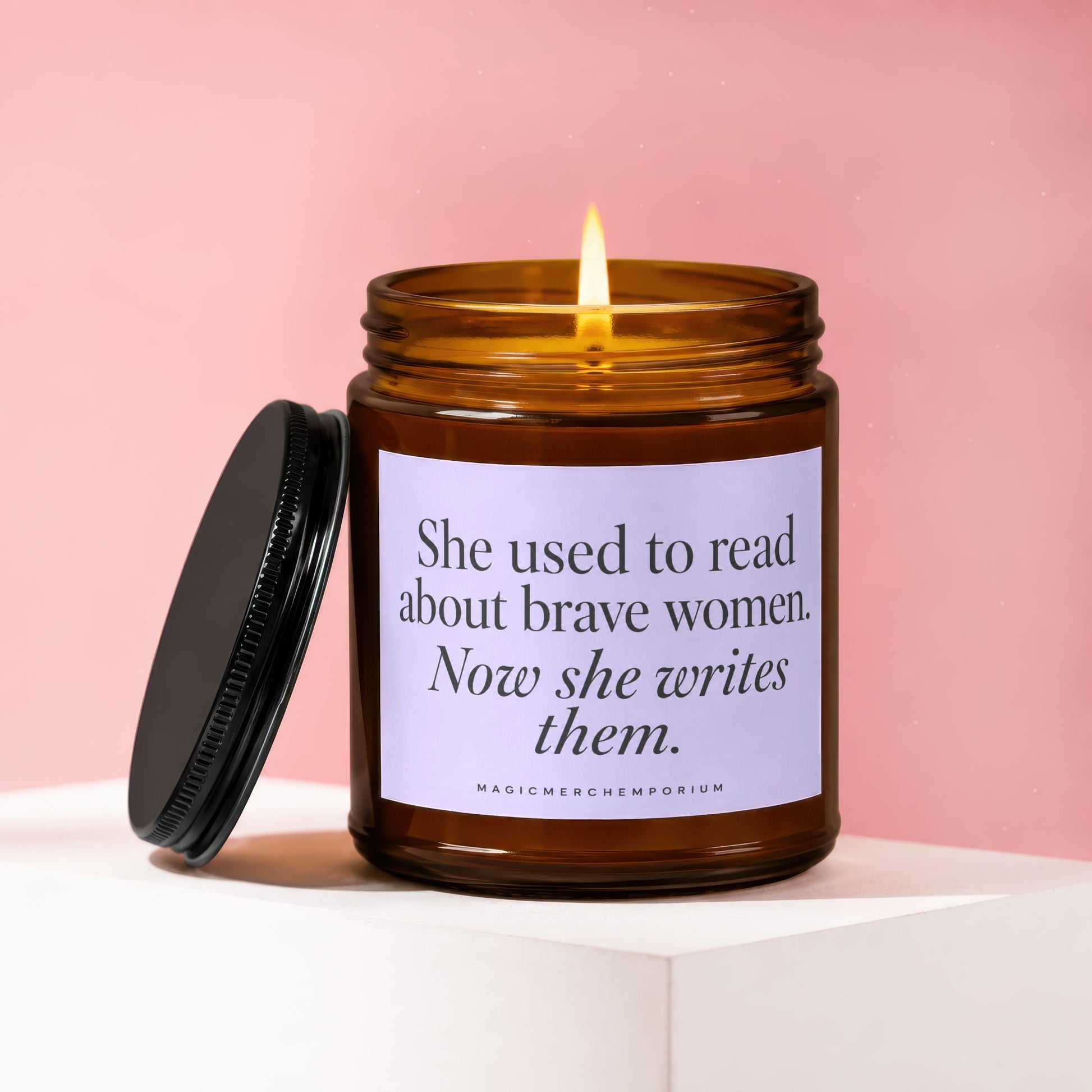 Amber glass candle with lit flame and text "She used to read about brave women. Now she writes them."