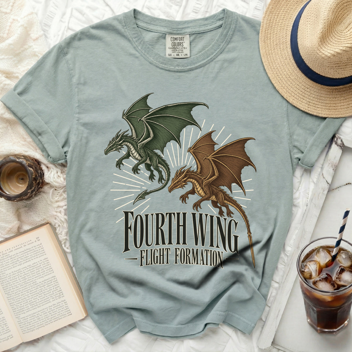 Sage green t-shirt featuring a green and brown dragon graphic and Fourth Wing Flight Formation text, surrounded by a book, hat, and iced drink.