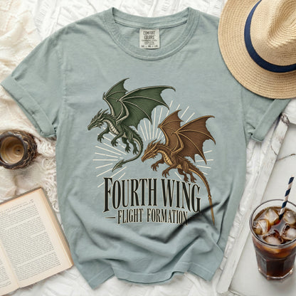 Sage green t-shirt featuring a green and brown dragon graphic and Fourth Wing Flight Formation text, surrounded by a book, hat, and iced drink.
