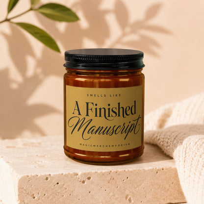 Candle with gold label reading "Smells Like A Finished Manuscript" and "MagicMerchEmporium."