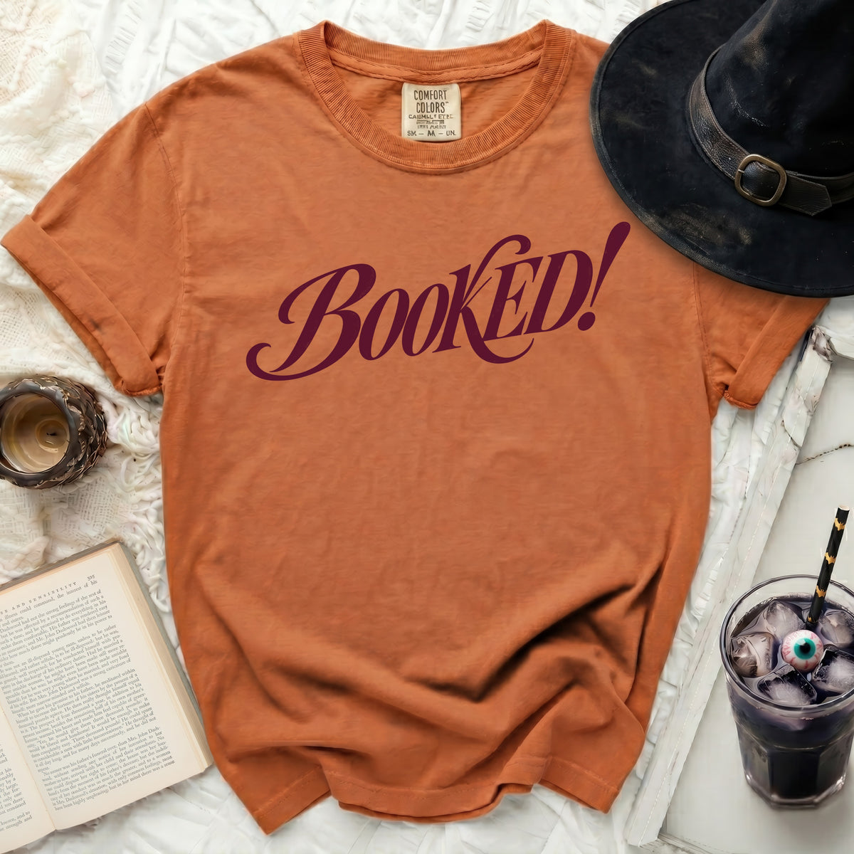 Orange t-shirt with "BOOKED!" text, witch hat, book, and drink.