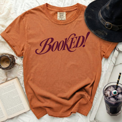 Orange t-shirt with "BOOKED!" text, witch hat, book, and drink.