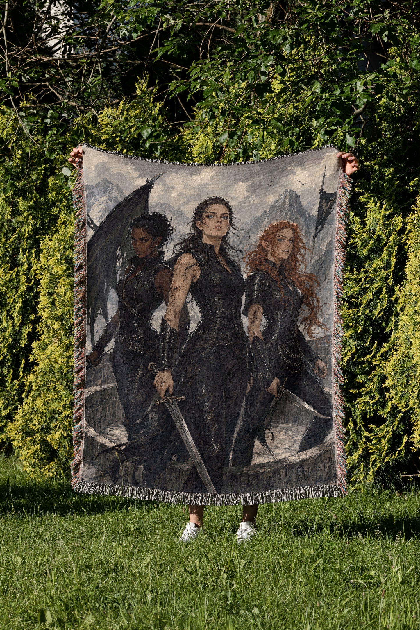 A person is holding up a large tapestry or blanket featuring three women in dark clothing, each holding a sword, in a grassy outdoor setting with trees in the background.